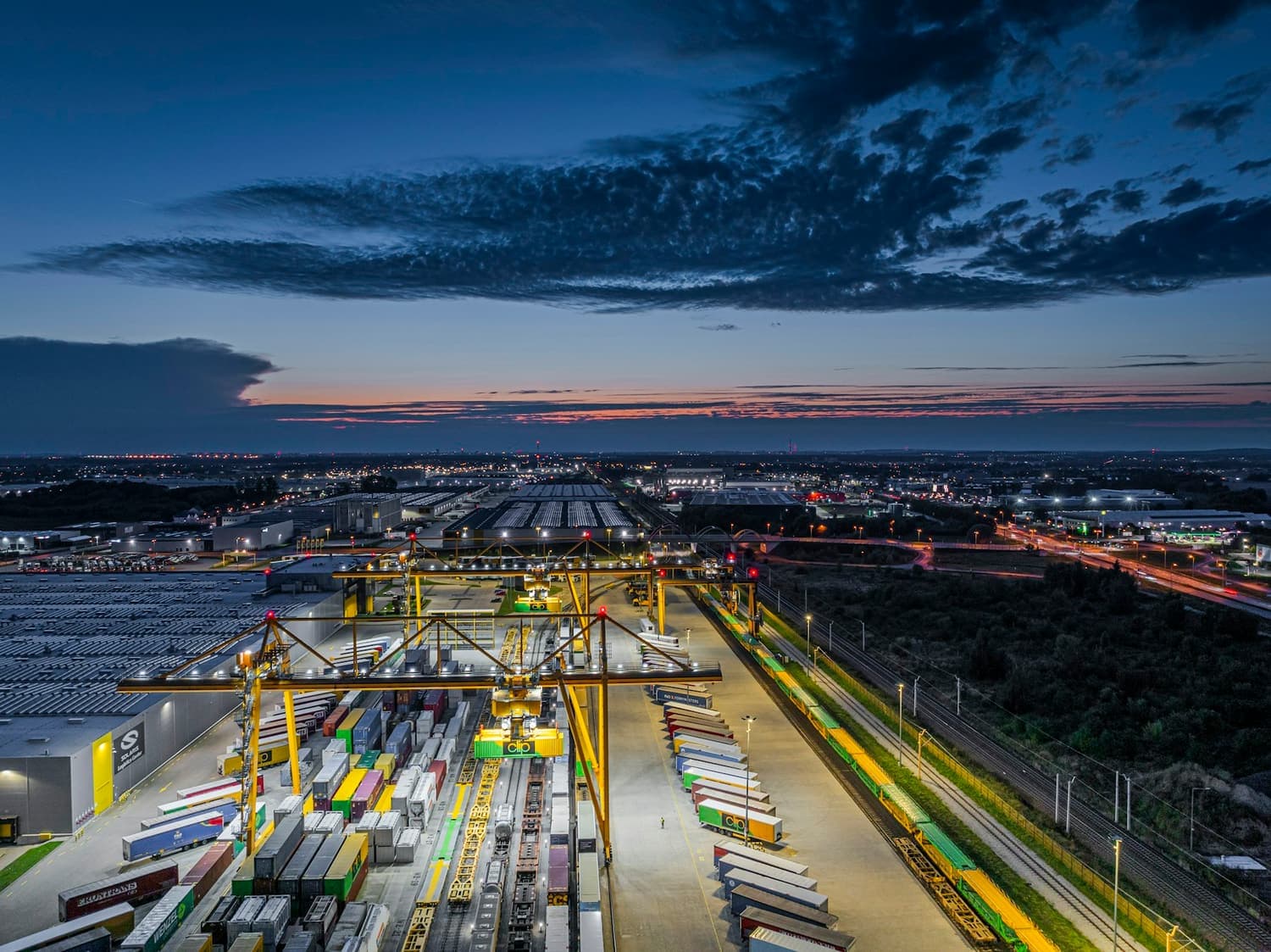 A container terminal at dusk, with gantry cranes and lines of waiting trailers